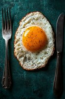 Sunny side up fried egg seasoned with black pepper on dark turquoise background, served with vintage fork and knife, dramatic food photography.