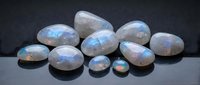Collection of polished rainbow moonstone gems displaying iridescent blue flash on dark surface for jewelry making and gemstone crafts.