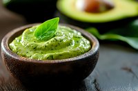 Fresh avocado sauce with basil leaf in wooden bowl, creamy green dip on dark background, half avocado blurred. Healthy vegan food concept.