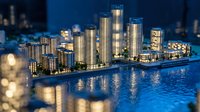 Illuminated architectural scale model of modern waterfront skyscrapers and residential buildings displayed at night with blue bokeh background.