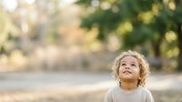 Toddler with curly blonde hair looking upward outdoors in natural sunlight, suitable for parenting, childhood, and lifestyle marketing materials.