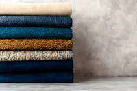 Stack of folded fabric swatches in navy, teal, mustard, and beige tones against a textured gray wall for interior design.