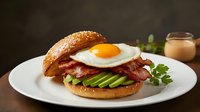 Gourmet breakfast burger with fried egg, crispy bacon and fresh avocado slices on sesame seed bun served on white plate for restaurant menu design.