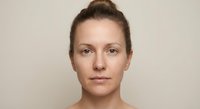 Natural portrait of young Caucasian woman with brown hair in bun, bare shoulders, neutral expression against beige background for beauty and skincare concepts.