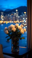 White roses in glass vase against evening cityscape with blurred bokeh lights reflecting on water, creating romantic atmosphere at twilight hour.
