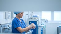 Female surgeon in blue scrubs and surgical cap preparing for operation in modern hospital operating room with medical equipment and bright lighting.