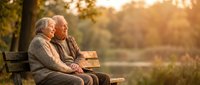 Elderly couple sitting together on park bench during golden hour sunset, enjoying peaceful retirement moment in nature surrounded by warm autumn light.