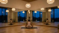 Serene yoga studio with warm pendant lighting and floor-to-ceiling windows overlooking twilight landscape. People practicing meditation on wooden floors.