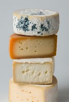 Stack of artisanal cheese wheels including blue cheese, aged cheddar, and soft ripened varieties on neutral background, showcasing different textures and colors.