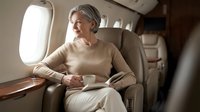 Elegant senior Caucasian businesswoman enjoying coffee while traveling in luxury private jet cabin, looking through window during flight.