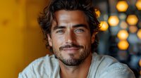 Confident Mediterranean man with dark curly hair and well-groomed beard smiling warmly against blurred bokeh lights background in casual attire.