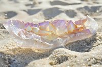 Translucent amethyst crystal bowl filled with colorful gemstones resting on sandy beach with soft natural lighting and ocean waves in background.