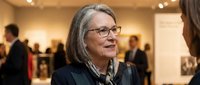 Senior businesswoman with gray hair and glasses smiling during professional networking event at modern gallery space with colleagues in background.