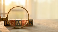 Magnifying glass examining wooden alphabet blocks spelling LEARN on desk with warm sunlight streaming through window for educational concept.