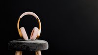 Pink and gold wireless headphones displayed on round gray cushion against dark background, minimalist product photography highlighting modern audio equipment.
