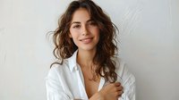 Young Caucasian woman with wavy brown hair wearing white button-up shirt smiling confidently against light background. Natural makeup and casual elegant style for business or lifestyle content.