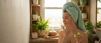 Young woman with towel on head applying skincare routine with cucumber slices in bright bathroom surrounded by green plants for natural beauty and wellness.