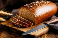Freshly baked whole grain bread loaf with sesame and poppy seeds on top, sliced on wooden cutting board with bread knife and wheat stalks.