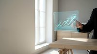 Businessman in dark suit touching transparent digital screen displaying upward trending arrow graph at wooden desk with natural window light, representing financial growth and business success.