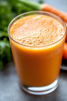 Fresh carrot juice with tiny bubbles in glass, bright orange color and blurred green herbs background, macro shot showing smooth texture and natural freshness.