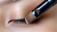 Professional makeup brush applying shimmering eyeshadow on closed eyelid, showing precise application technique and soft neutral tones for cosmetic artistry.