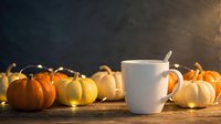 Blank white mug surrounded by decorative mini pumpkins and warm string lights on wooden surface, autumn seasonal beverage mockup.