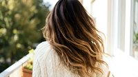 Young woman with long wavy brown ombre hair in cream cable knit sweater relaxing on sunny balcony with natural lighting and soft focus background.