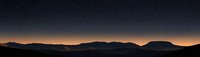 Panoramic mountain silhouettes against dark starry night sky with glowing orange horizon line creating dramatic natural landscape at dusk.