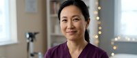 Mature Asian woman wearing purple scrubs smiles at camera in medical clinic setting with warm lighting and blurred background. Healthcare professional portrait.