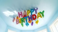 Colorful foil letter balloons spelling Happy Birthday float against a light blue ceiling with white balloons, festive party decoration.