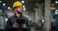 Industrial worker in yellow hard hat using digital tablet in modern factory warehouse for safety inspection and quality control operations.