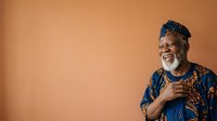 Mature African male with gray beard wearing traditional blue and orange patterned clothing, smiling warmly against warm tan background for portrait and lifestyle use.