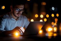 Young professional man with glasses working late on laptop computer in dark office with warm bokeh lights creating atmospheric nighttime workspace ambiance.