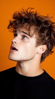 Young Caucasian male model with messy brown hair looking up expressively against vibrant orange background, wearing black crew neck sweater. Studio portrait capturing genuine emotion.