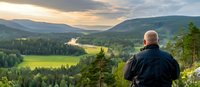 Senior male photographer capturing panoramic mountain valley view at sunset, with winding river and green forests stretching to distant hills under dramatic sky.