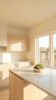 Bright, minimalist kitchen with light wood cabinetry, white countertops, and large windows flooding the space with natural sunlight. Potted succulent plant on island. Modern residential interior design.
