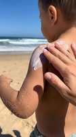 Young boy applying sunscreen lotion to his shoulder at the beach on a sunny day, protecting skin from UV rays during summer vacation and outdoor activities.