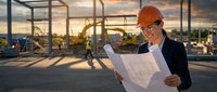 Professional female construction engineer reviewing architectural blueprints at building site during golden hour sunset with steel framework background.