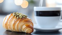 Fresh baked flaky croissant topped with crushed pistachios served with black coffee in white cup against blurred bokeh background for cafe menu.