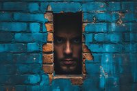 Man peering through rectangular hole in vibrant turquoise brick wall, creating dramatic contrast between shadow and light.