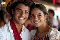 Young Hispanic couple in traditional Mexican folk costumes smiling at camera during cultural festival celebration with colorful decorations in background.