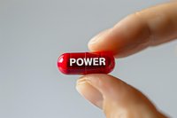 Red capsule pill with power text held between fingers against light background, representing strength and energy boost concept for health supplements.