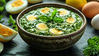 Creamy spinach soup with halved boiled eggs in rustic ceramic bowl, garnished with fresh herbs and sesame seeds on dark wooden surface.