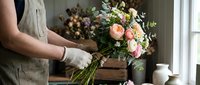 Female florist arranging fresh pink roses and mixed flowers in rustic workshop setting. Professional flower arrangement and bouquet making for weddings.