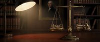 Brass scales of justice on wooden desk with overhead lamp in law office library setting with books and dark background.