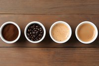 Four white bowls on wooden table showing coffee stages: ground powder, roasted beans, espresso with foam, and latte with milk, top view.
