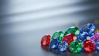 Colorful precious gemstones collection with emerald, ruby, sapphire and diamond stones scattered on reflective surface for luxury jewelry design.