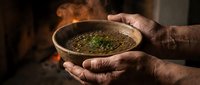 Hands holding steaming bowl of soup with herbs over fire, rustic cooking scene. Warm comfort food preparation.