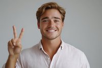 Young Caucasian man with brown hair wearing pink button-down shirt showing peace sign gesture against gray background, expressing positive cheerful emotion.
