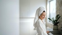 Young Caucasian woman in white bathrobe with towel on head relaxing in modern bathroom after shower. Morning skincare routine and wellness concept.